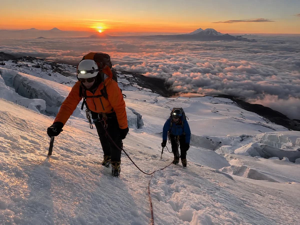 Dos andinistas encordados ascienden por un glaciar al amanecer, sobre un mar de nubes.