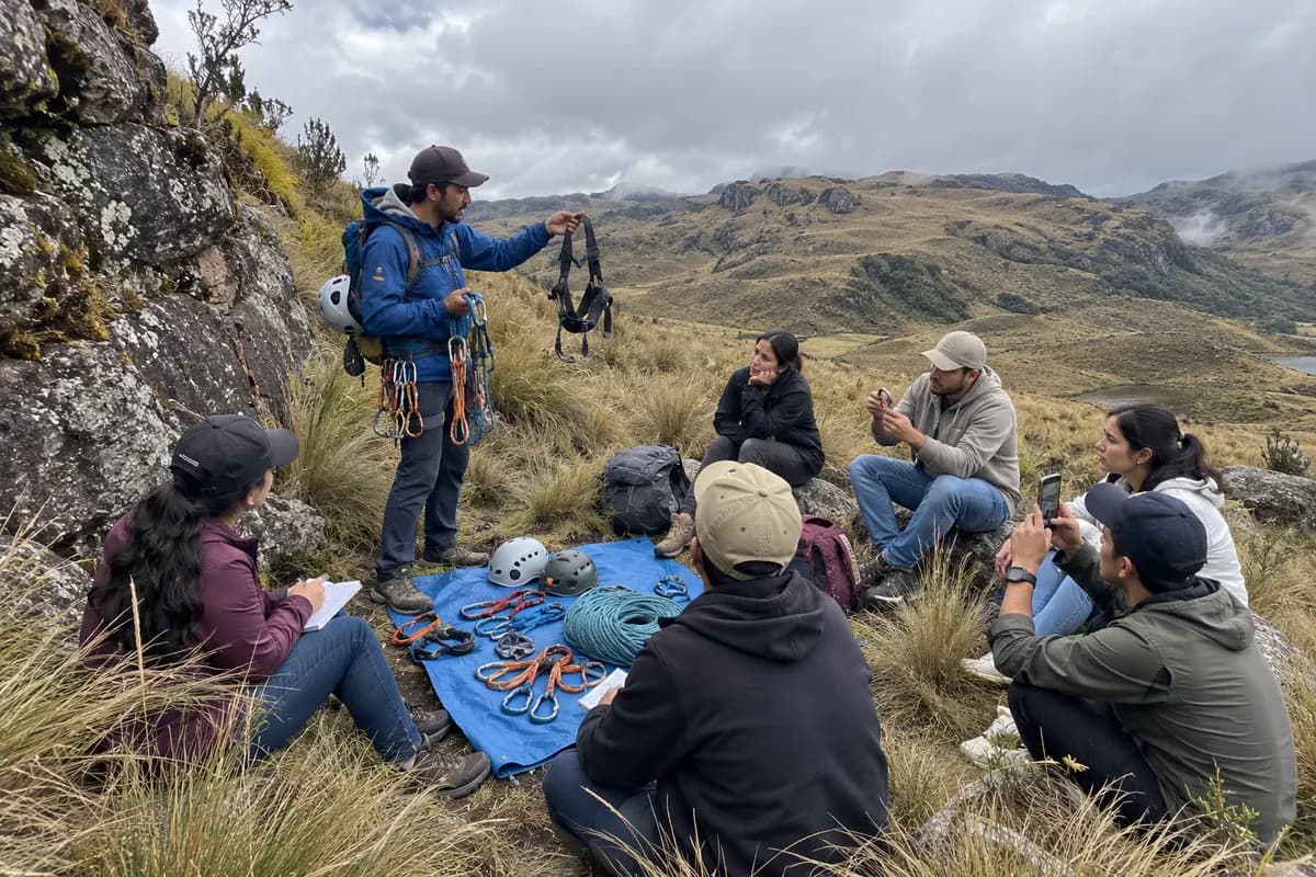 Instructor muestra arneses y mosquetones a un grupo sentado en el páramo, con el equipo de escalada sobre una lona.