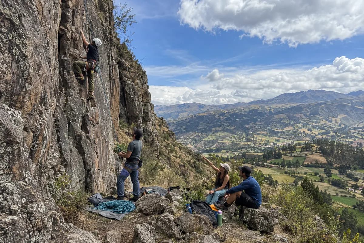 Escalador asegurado en una pared de roca mientras sus compañeros lo miran desde abajo, con un valle andino al fondo.